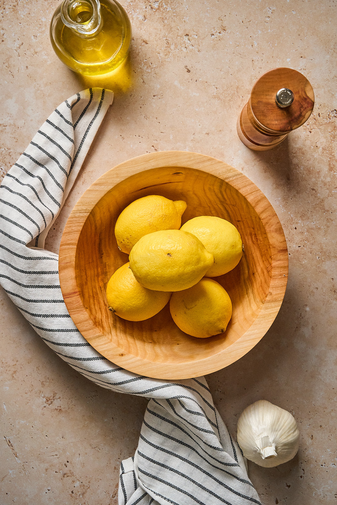 Handmade wooden bowl filled with fresh lemons, next to garlic, olive oil, and a striped towel on a countertop.