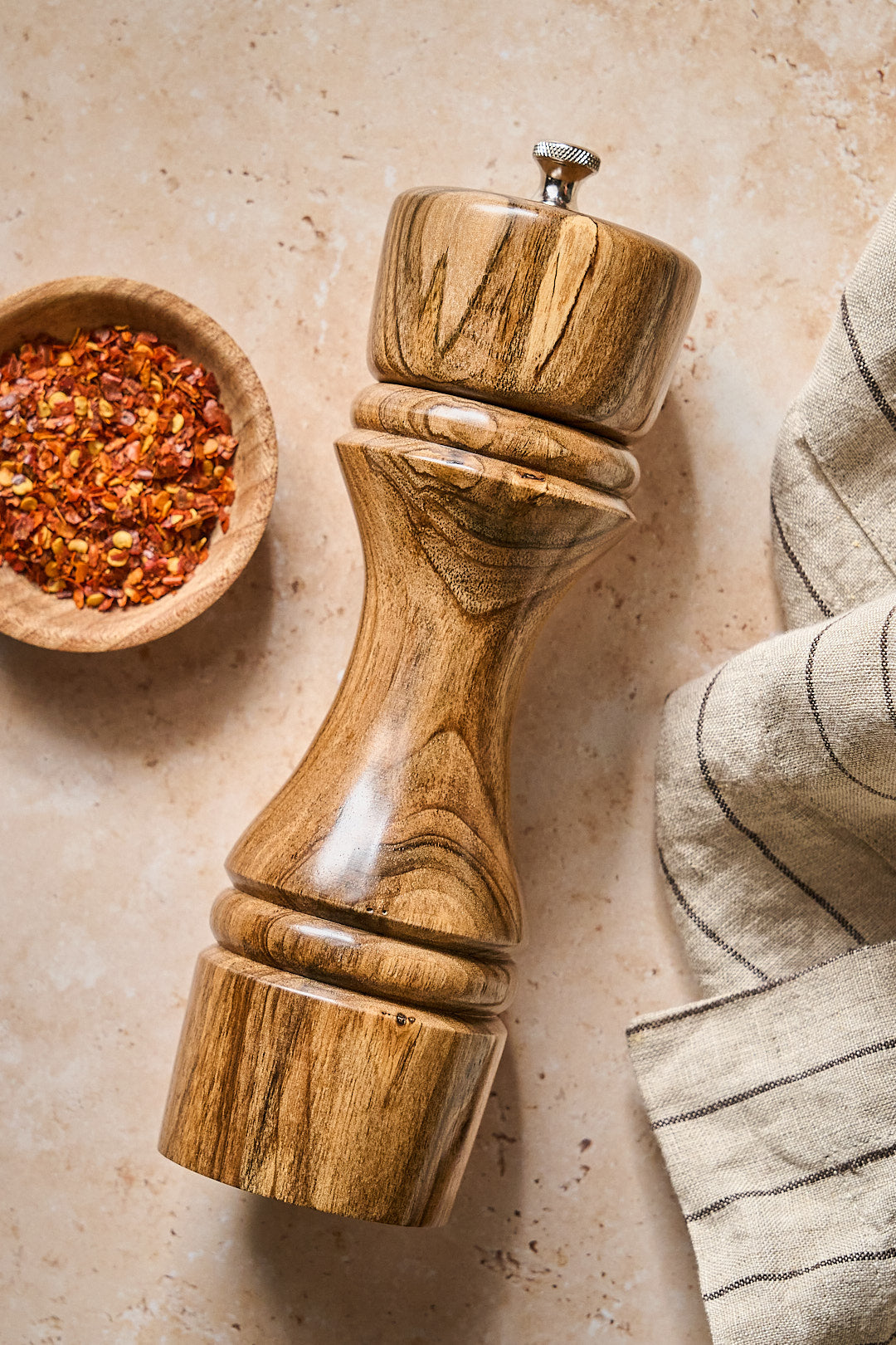 Handmade walnut pepper grinder beside a bowl of spices and a striped kitchen towel.