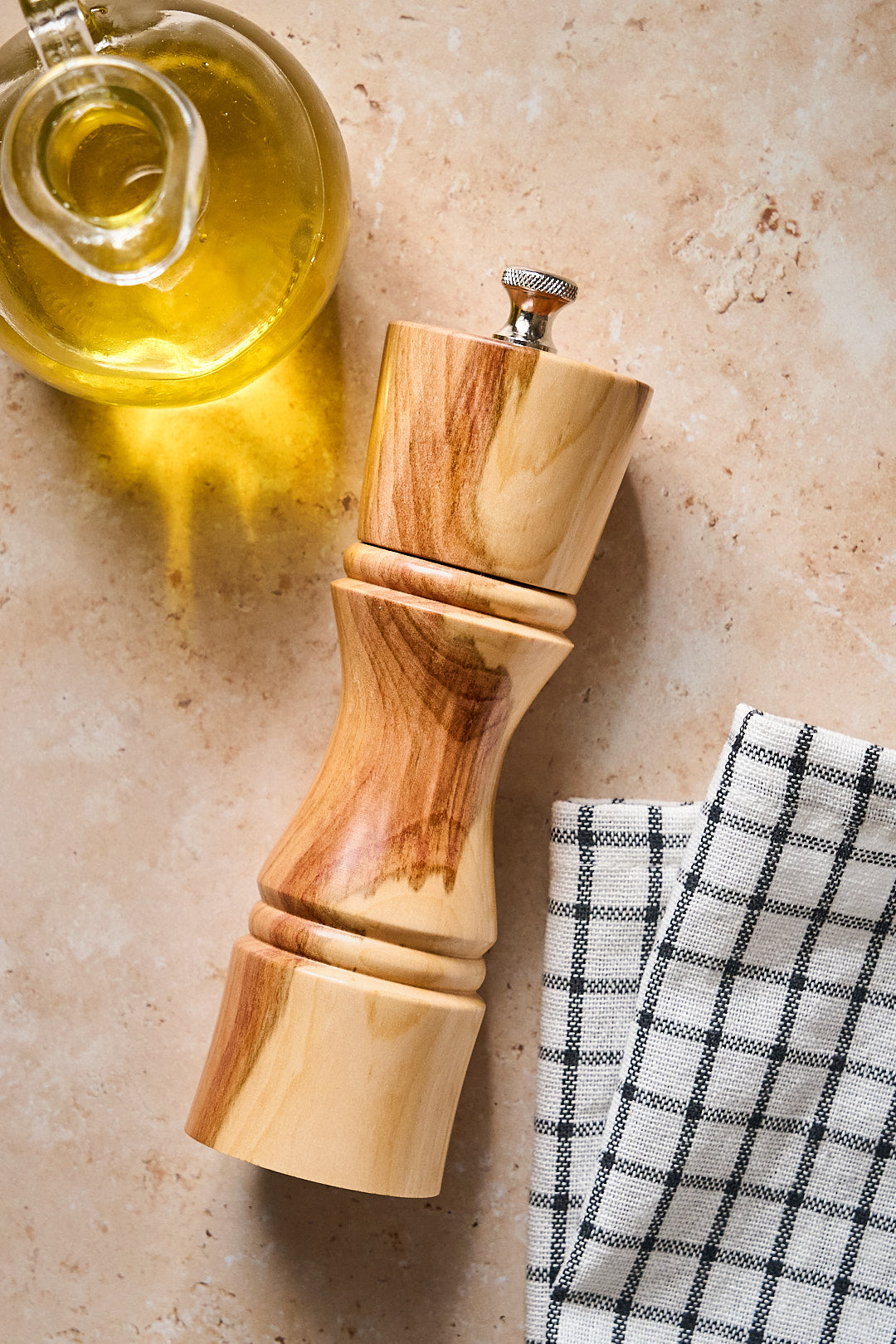 Overhead view of small handmade pepper mill next to a checkered linen and a bottle of oil