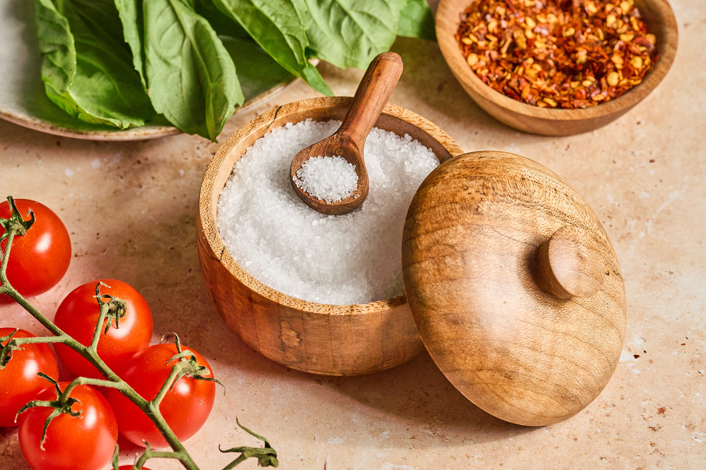 Maple wood salt cellar next to tomatoes, herbs, and spices on a stone background