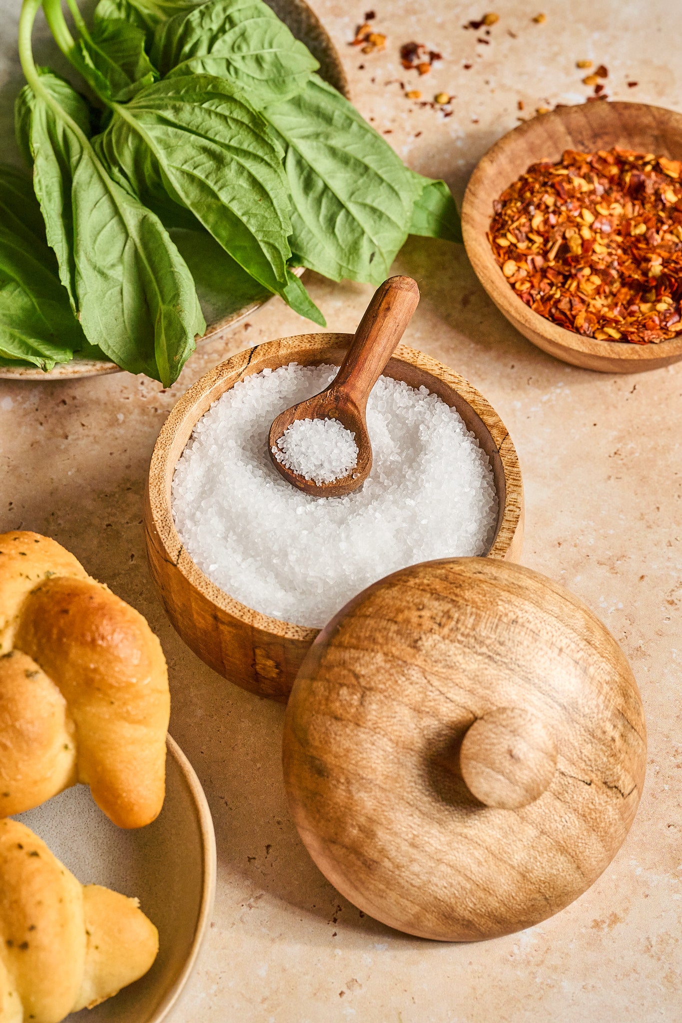 The wood salt cellar with a tiny spoon resting inside next to herbs, spices, and bread.